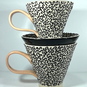 May include: Two stacked ceramic coffee cups with a black and white squiggly pattern. The cups have peach-colored handles and sit on a black saucer. The cups are a cream color.