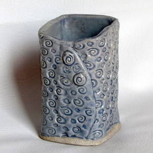May include: A light blue ceramic vase with a swirling pattern. The vase has a rectangular shape and a slightly uneven base.