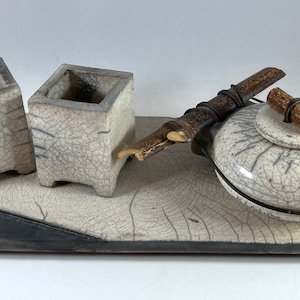 May include: A white ceramic tea set with a teapot, two small square containers, and a wooden handle. The set is displayed on a black and white ceramic tray.