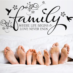 Könnte beinhalten: Ein schwarz-weißes Wandtattoo mit dem Wort "Familie" in einer kursiv geschriebenen Schrift. Das Tattoo enthält außerdem ein florales Design und zwei Kolibris. Der Text unter dem Wort "Familie" lautet "Wo das Leben beginnt & die Liebe nie endet". Das Tattoo ist an einer weißen Wand über einem Bett angebracht, aus dem fünf Füße aus den Decken ragen.