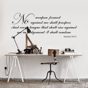 Peut inclure: Un mur blanc avec une citation inspirante en noir et blanc qui dit "No weapon formed against me shall prosper; And every tongue that shall rise against me in judgement I shall condemn ISAIAH 54:17". La citation est affichée au-dessus d'un bureau blanc avec une lampe noire, un appareil photo noir et une chaise en bois.