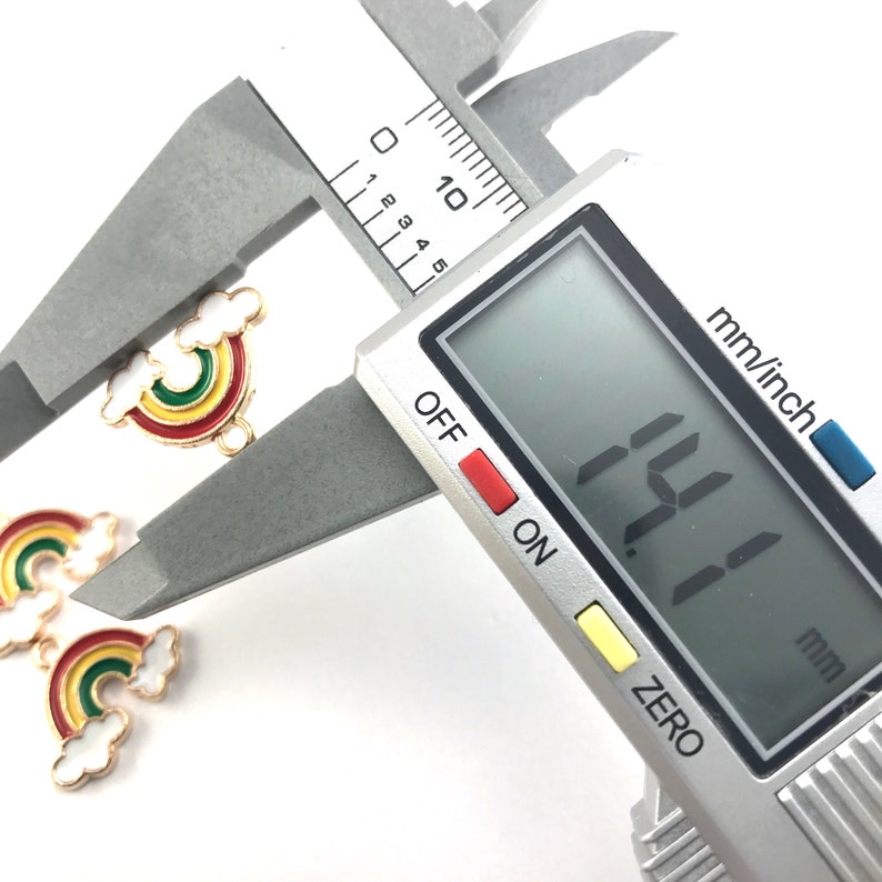 May include: Close-up of a rainbow charm with a cloud detail, measured with a digital caliper showing 1.11 inches. The charm is gold-toned with red, yellow, green, and blue enamel.