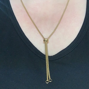 May include: A gold-coloured necklace featuring a fine chain and a sliding pendant. The pendant has a dangling chain with two small, round charms. The necklace is worn against a black top.
