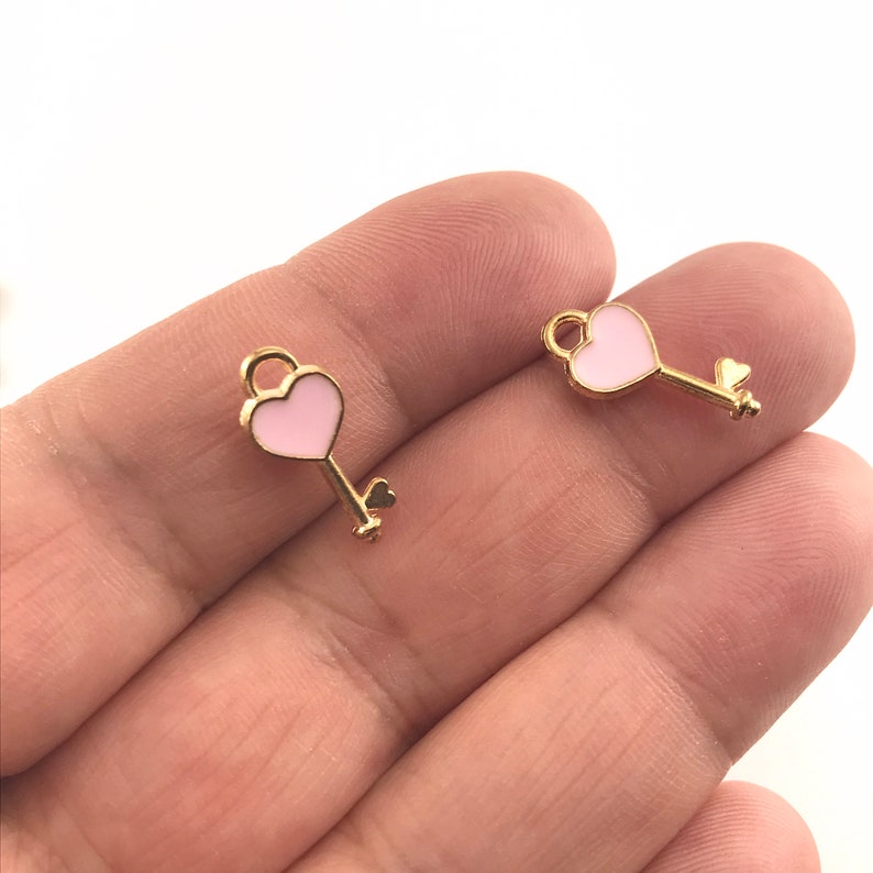 May include: Two gold-colored heart-shaped key charms with pink enamel. The charms have a small loop at the top for attaching to a necklace or bracelet.