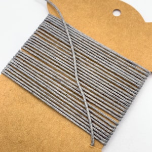 May include: A spool of grey twine wound around a brown cardboard card. The twine is made of natural fibres and has a rustic look.