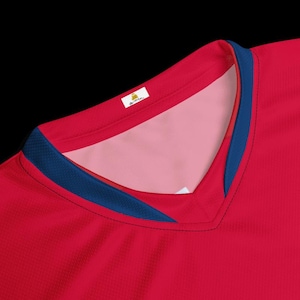 May include: A red V-neck jersey featuring a dark blue trim along the collar. The interior of the neckline is a light pink color. A small white rectangular tag with black text is visible on the inner back collar of the athletic shirt.