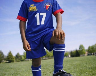 May include: A young person in a blue soccer uniform with red trim and the number 17. The uniform features a crest and the word "HAITI". They are standing on a soccer ball, wearing black cleats and blue socks with white stripes, on a grassy field.