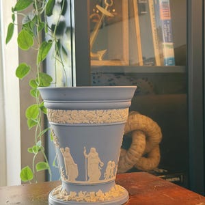May include: A light blue ceramic vase featuring white relief ornamentation. The design includes a band of floral motifs at the top and bottom, with a central frieze depicting classical figures and cherubs in a raised, matte white finish.