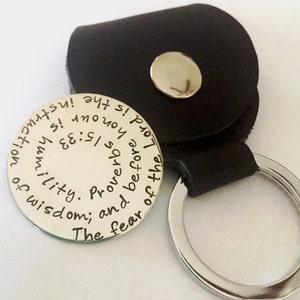 Op de afbeelding: Een zilveren sleutelhanger met een zwart leren bandje. De sleutelhanger heeft een ronde metalen schijf met de inscriptie "The fear of the Lord is the beginning of wisdom; and Proverbs 9:10 Humility is instruction!"