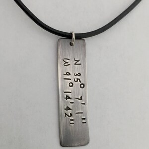 May include: A silver necklace with a rectangular pendant featuring the coordinates "N 35° 7' 1" W 91° 14' 42" ". The necklace is on a black cord.