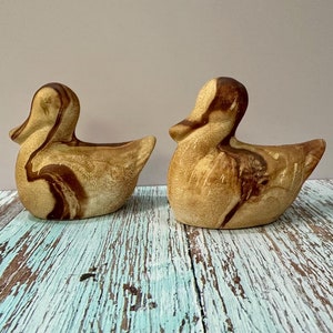 May include: Two ceramic duck figurines with brown and tan marbling. The ducks are sitting on a light blue and brown wooden surface.