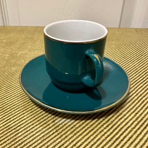 May include: Teal-colored teacup and saucer set with gold trim. The teacup has a white interior and a curved handle. The saucer is round and matches the cup's color. The set is made of ceramic and is suitable for serving tea or coffee.