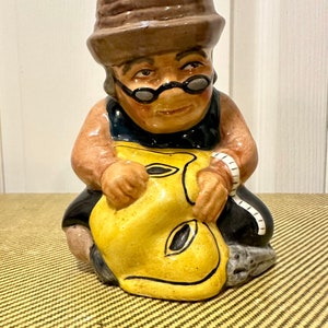 May include: A ceramic figurine of a person wearing a brown hat and glasses, holding a yellow object with black details. The figurine is sitting on a textured surface.