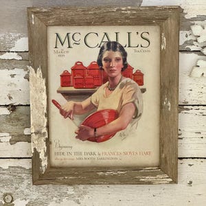 May include: Framed vintage McCall's magazine cover from March 1929. The artwork features a woman in a yellow dress holding a red bowl and spoon. Red spice jars are on a shelf behind her. The frame is rustic with distressed edges.