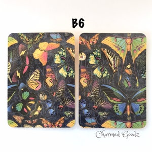 May include: Two rectangular coasters with rounded corners. The coasters feature a dark background covered in colorful butterflies. The text "B6" is at the top, and "Charmed Goodz" is at the bottom. The butterflies are in shades of yellow, pink, blue, and green.