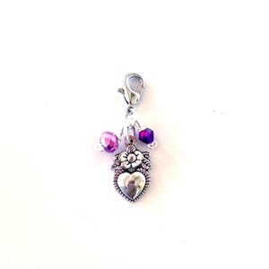 May include: A silver-toned charm with a heart-shaped pendant, adorned with a flower and leaf design. The charm features a lobster clasp and is embellished with purple and pink beads, creating a decorative accessory.