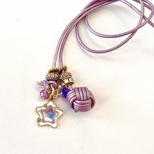 May include: A lavender leather cord necklace featuring a knotted leather accent, gold-toned beads, and a dangling star-shaped charm with a pearlescent center, white enamel border, and small embedded crystals.