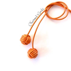 May include: Two knotted leather tassels in a burnt orange color. The tassels are connected by a long leather cord.