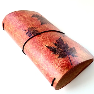 May include: A brown leather journal with a red and orange marbled pattern and black leaf designs. The journal is closed and has a black elastic band around it.