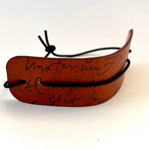 May include: A brown leather bracelet with black elastic bands. The leather is curved and features handwritten text in black ink. The bracelet is designed to wrap around the wrist and is set against a white background.