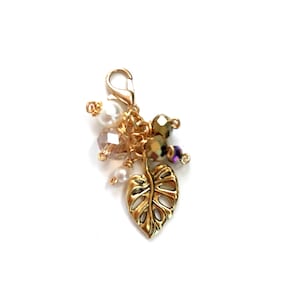 May include: A gold-colored charm with a lobster clasp, featuring a leaf pendant and clusters of beads in various colors. The beads include pearls, clear, and iridescent beads. The charm is designed for jewelry making or as a decorative accessory.