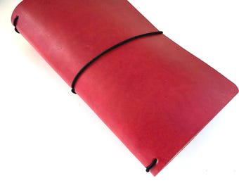 May include: A red leather journal with a black elastic closure. The journal is closed and has rounded corners. The cover is a solid, vibrant red color. The journal is lying on a white surface.