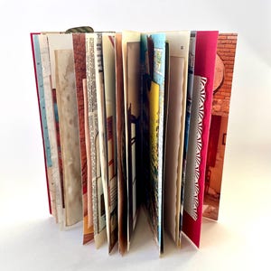May include: An open journal with a red cover and a variety of pages. The pages include printed text, illustrations, and decorative elements. The journal is standing upright, showcasing the different textures and colors of the pages.