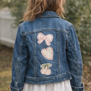May include: Blue denim jacket with pink bow, heart, and cherry patches on the back. The jacket has a classic collar, button-up front, and long sleeves. The patches are stitched onto the denim.