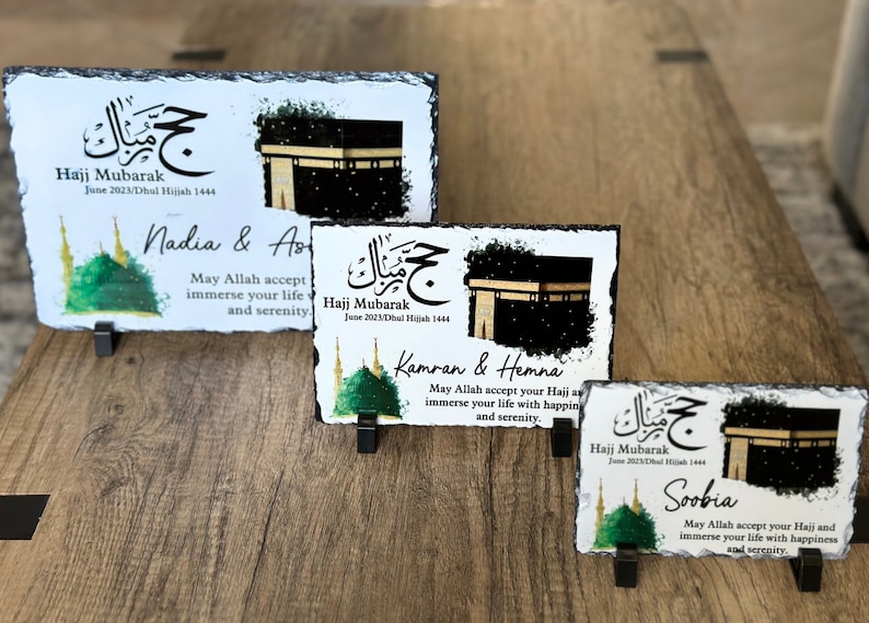 May include: Four slate plaques with a white background and black borders. Each plaque features a green watercolor image of a mosque, a black and white image of the Kaaba, and Arabic calligraphy. The text on the plaques reads "Hajj Mubarak" and includes the date. The plaques also include personalized names and a message wishing the recipients a blessed Hajj.