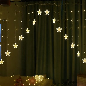May include: String lights with warm white LED lights shaped like stars. The star lights are hanging from a wire and are in front of a dark green curtain. The lights are on, illuminating the room.