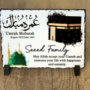 May include: A white slate plaque with a black and gold image of the Kaaba, a green mosque, and Arabic calligraphy. The text reads "Umrah Mubarak August 2023/Safar 1445 Saeed Family May Allah accept your Umrah and immerse your life with happiness and serenity."