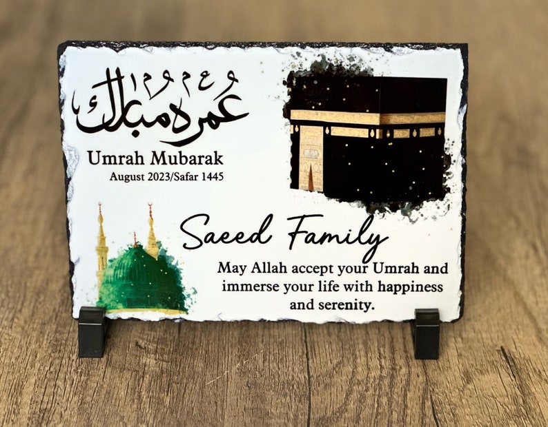 May include: A white slate plaque with a black and gold image of the Kaaba, a green mosque, and Arabic calligraphy. The text reads "Umrah Mubarak August 2023/Safar 1445 Saeed Family May Allah accept your Umrah and immerse your life with happiness and serenity."