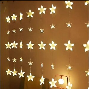 May include: A string of warm white LED lights shaped like stars, hanging from a ceiling. The lights are arranged in a cascading pattern, creating a festive and whimsical atmosphere.