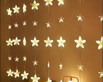 May include: A string of warm white LED lights shaped like stars, hanging from a ceiling. The lights are arranged in a cascading pattern, creating a festive and whimsical atmosphere.