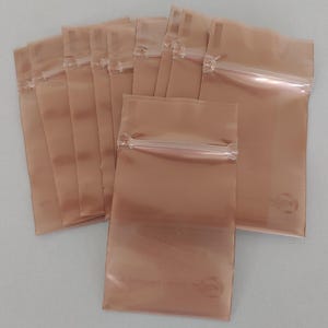 以下が含まれることがあります： 銅色の再封可能なプラスチックバッグのコレクション。各バッグには透明なジッパー式の開口部があり、右下の角には小さな円形のロゴが付いています。バッグは扇状に配置されており、一部が重なっています。