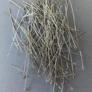 Op de afbeelding: Een stapel zilverkleurige kopspelden met lusvormige uiteinden, verspreid op een grijze ondergrond. De pinnen zijn dun en recht, met een kleine lus aan één uiteinde, voor het maken van sieraden en knutselen.