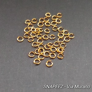 May include: A pile of gold jump rings, a type of jewelry finding used for connecting chains, cords, and other jewelry components.  The jump rings are small and round, and they are scattered on a gray surface.