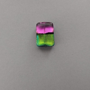 May include: A rectangular glass bead with a dichroic finish. The top half is a vibrant pink, transitioning to a black band, then a bright green on the bottom. The bead is likely for jewelry making.
