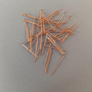 Op de afbeelding: Een verzameling koperkleurige spelden verspreid op een grijze ondergrond. De spelden hebben kleine, ronde koppen en lange, slanke schachten. De spelden zijn in een willekeurig patroon gerangschikt.