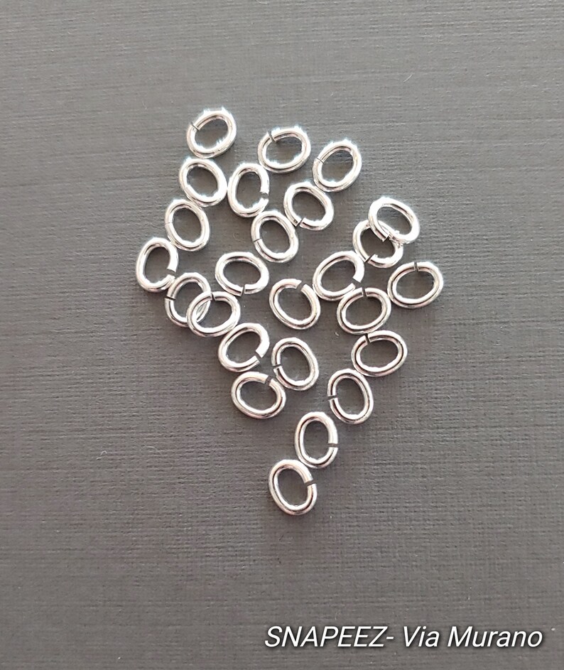 May include: A pile of silver jump rings, oval shaped, on a gray background.  SNAPEEZ - Via Murano.