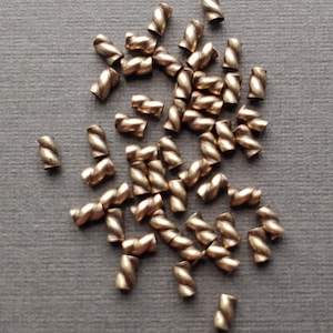 May include: A pile of gold-colored metal crimp beads with a twisted design. The beads are scattered on a gray surface.