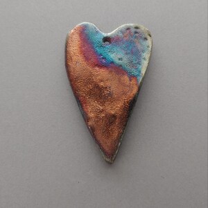 May include: A heart-shaped pendant featuring a textured surface with a metallic copper finish that transitions into iridescent blue and purple hues. The piece includes a small circular hole at the top for suspension, displaying a weathered, uneven edge profile.