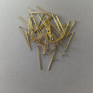 Op de afbeelding: Een verzameling goudkleurige kopspelden verspreid op een grijze ondergrond. De spelden hebben een kleine, ronde kop en een lange, slanke schacht. Ze worden waarschijnlijk gebruikt voor naaien, knutselen of het maken van sieraden.