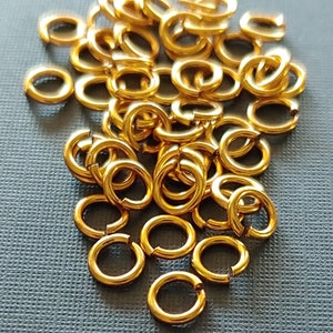 May include: A pile of gold-colored jump rings, open and closed, scattered on a dark gray surface. The rings are shiny and metallic, with a circular shape. The text "SNAPEEZ - Via Murano" is at the bottom.