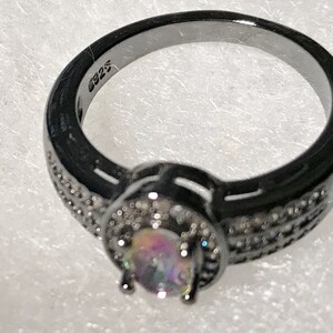 May include: Black ring with a round iridescent gemstone surrounded by smaller clear gemstones. The ring is marked with "925" on the inside band.
