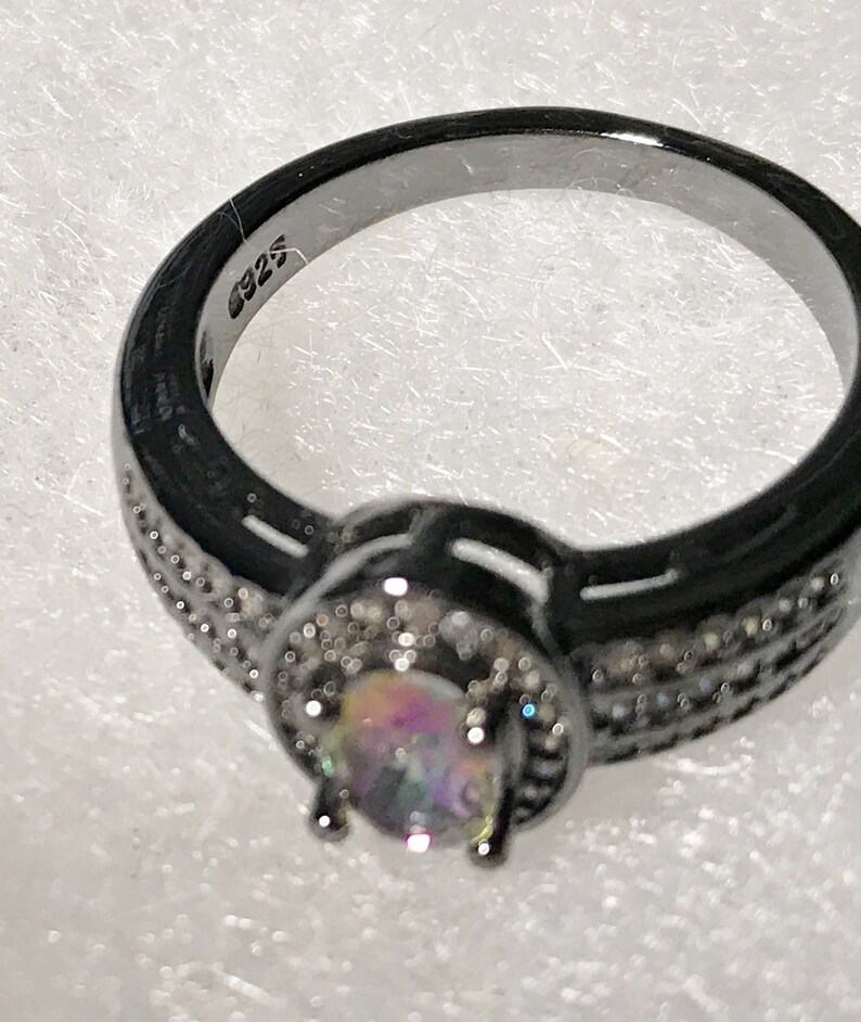 May include: Black ring with a round iridescent gemstone surrounded by smaller clear gemstones. The ring is marked with "925" on the inside band.