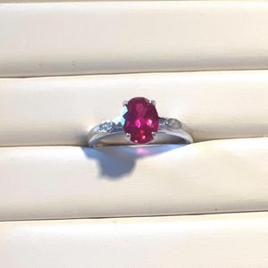 May include: A silver ring featuring a large, oval-cut, vibrant red gemstone as the centerpiece. The ring is flanked by smaller, clear accent stones on each side. The setting is simple and elegant, showcasing the brilliance of the gemstones.