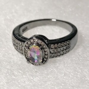 May include: A black ring with a large oval-shaped iridescent gemstone surrounded by smaller clear gemstones. The ring has a black band with a double row of smaller clear gemstones.