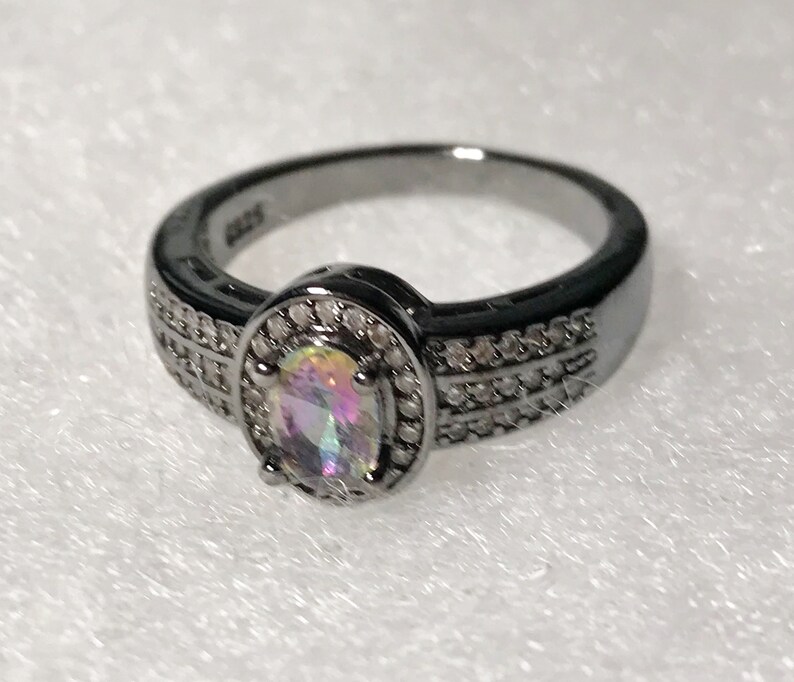May include: A black ring with a large oval-shaped iridescent gemstone surrounded by smaller clear gemstones. The ring has a black band with a double row of smaller clear gemstones.