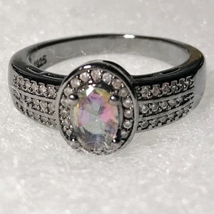 May include: A black ring with a large oval-shaped iridescent gemstone surrounded by a halo of small clear stones. The band of the ring is decorated with two rows of small clear stones.